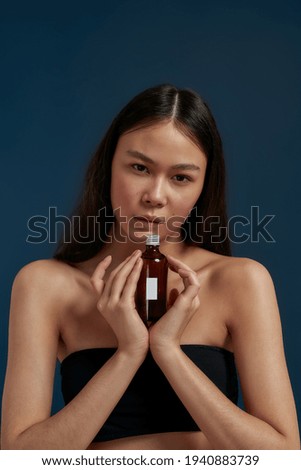 Asian model looking at the camera while holding a personal care product. Stylish and well-grooming girl standing on a dark background. Girl in a black top with a personal care product