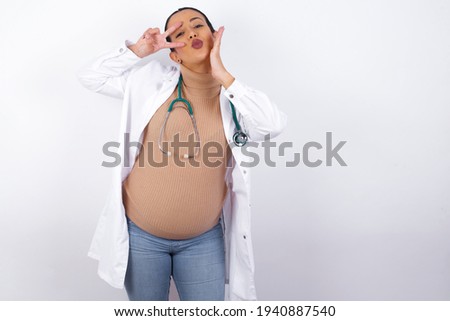 young pregnant doctor woman wearing medical uniform against white background  making v-sign near eyes. Leisure lifestyle people person celebrate flirt coquettish concept.
