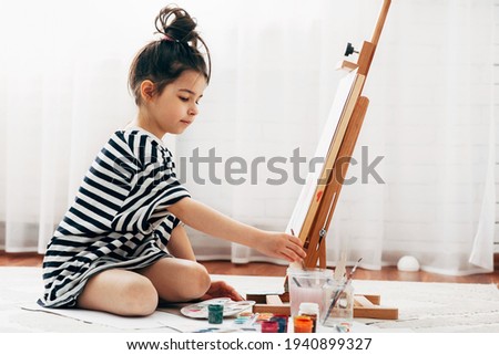 Horizontal full-length image of a little girl kid painting on paper on an easel at home. A cute kid sitting on the carpet and drawing in her room.