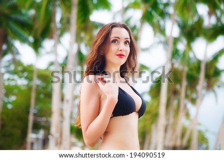 Beautiful young woman with long hair in black bikini relaxes under palm trees, sea in the background.