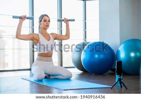 Female fitness blogger with dumbbells in hands records training video on smartphone camera