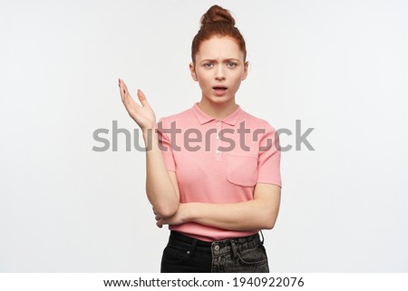 Unhappy lady, frowning woman with ginger hair bun. Wearing pink t-shirt and black jeans. Raise her hand in disagreement. Watching at the camera, isolated over white background.
