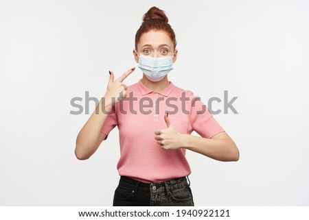 Portrait of girl with ginger hair bun. Wearing pink t-shirt and medical, protective face mask. Pointing at the mask and shows thumb up. Watching at the camera, isolated over white background