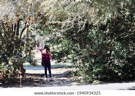 A young girl in a pink jacket walking under a bridge made of holly plants. The holly is blooming and has fresh, red berries throughout it. She's reaching to grab some berries while exploring nature.
