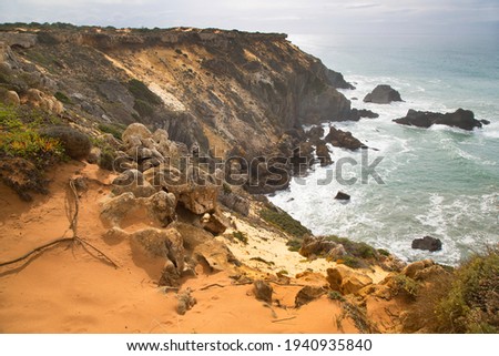 typical natural rugged coastline landscape of south west Portugal with sand dunes, rocks, undergrowth and pebbly beach with a threatening rain cloudy sky
