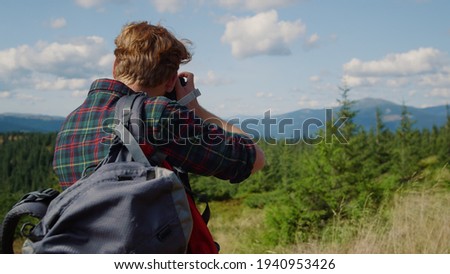 Back view male photographer using photo camera outdoor. Redhead man taking photos during hike in green mountains. Smiling guy checking pictures on photo camera. Young hipster enjoying vacation 