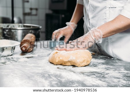 Female hands knead dough on the table next to a piece of dough and a baking dish. 