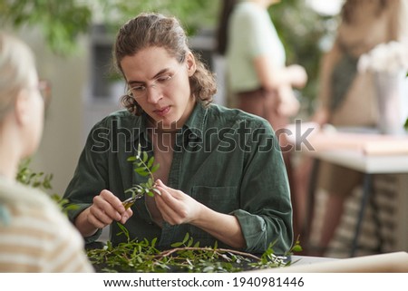 Portrait of young male florist cutting green plants while creating floral compositions for decor, copy space