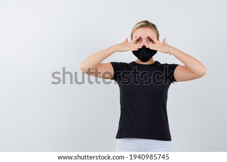  blonde lady looking through fingers in black t-shirt, black mask and looking curious, front view. 