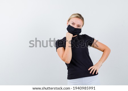 Portrait of blonde lady propping chin on hand in black t-shirt, black mask and looking thoughtful front view 