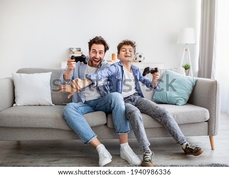 Joyful dad and son with joysticks playing video games at home, boy distracting father with hand
