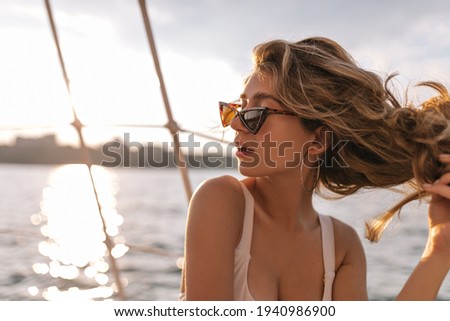 Young lady with fluffy blonde hair in modern beach outfit and dark sunglasses posing in view of sea at sunshine and looking away