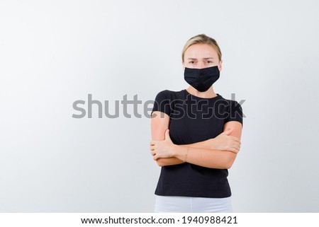 blonde lady standing with crossed arms in black t-shirt, black mask and looking confident, front view. 