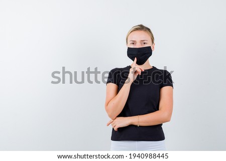  blonde lady holding finger on her medical mask in black t-shirt and looking confident. front view. 
