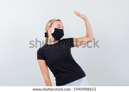 Portrait of blonde lady waving hand while closing eyes in black t-shirt, black mask and looking relaxed front view 