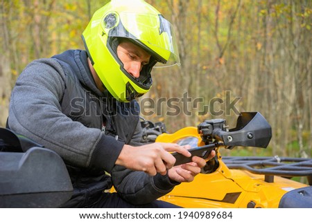 ATV driver holds a smartphone in his hands. Portrait of a motorcyclist with a phone. Concept - ATV driver it uses a navigation app. Navigation app for bikers. Biker on background of forest.