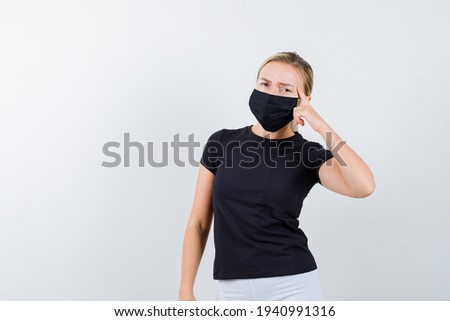  blonde lady pressing finger on temples in black t-shirt, black mask and looking serious. front view. 