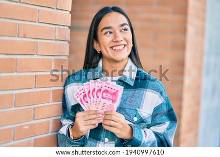 Young latin girl smiling happy holding chinese yuan banknotes at the city.