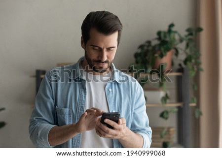 Close up focused man looking at phone screen, standing at home, browsing mobile device apps, reading news, chatting or shopping online, searching information in internet, holding smartphone Royalty-Free Stock Photo #1940999068