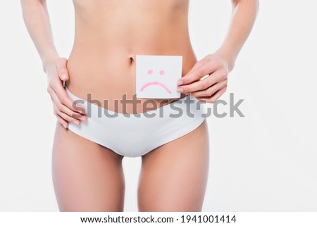Partial view of slim woman in panties holding card with sad emoticon sign isolated on white