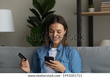 Close up smiling woman holding plastic card and phone, sitting on couch at home, happy young female making secure internet payment, satisfied customer shopping online, browsing banking service