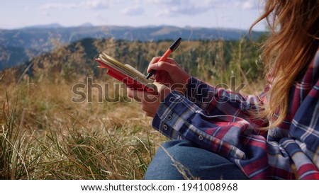 Talented girl hands creating sketch of trees in notebook. Attractive woman drawing picture with pen at weekend in mountains. Smiling female hiker sitting in field with notepad during hike 