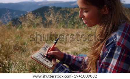 Happy woman drawing sketch of trees in notebook at weekend in mountains. Positive girl painting picture with pen. Smiling female hiker enjoying natural landscape. Young lady taking rest during hike