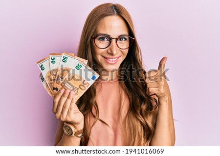 Young brunette woman holding bunch of 50 euro banknotes smiling happy and positive, thumb up doing excellent and approval sign 