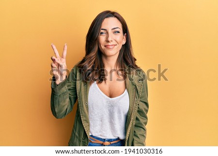 Young brunette woman wearing casual clothes over yellow background smiling with happy face winking at the camera doing victory sign. number two. 