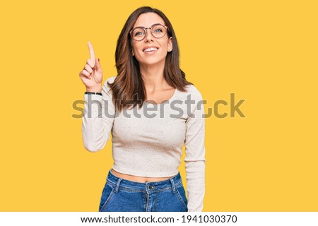 Young brunette woman wearing casual clothes and glasses showing and pointing up with finger number one while smiling confident and happy. 