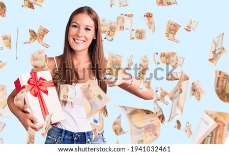 Young beautiful hispanic woman holding gift smiling happy and positive, thumb up doing excellent and approval sign
