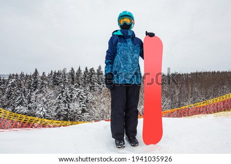 Female in a blue suit standing with a snowboard on a large snow slope. Snowboarder looks at a beautiful view of the snowy forest. Skiing holidays. Logoisk, Belarus