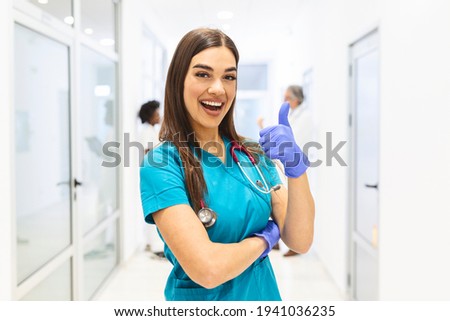 Portrait Of Smiling Female Doctor Wearing Scrubs In Busy Hospital Corridor showing thumbs up