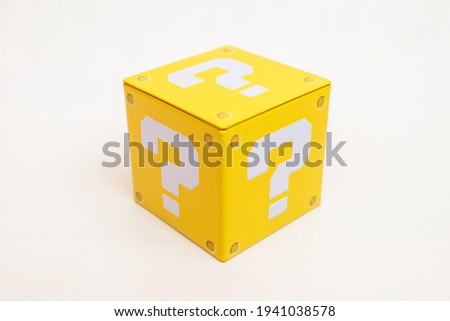 Indoor still-life photo of a yellow box with a big white question mark printed on each face. It recalls a graphic element of a famous platform video game. Royalty-Free Stock Photo #1941038578