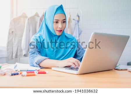 Asian Muslim woman fashion designer job occupation working in clothing textile workshop modern office researching design using computer laptop, designing planning using software application project