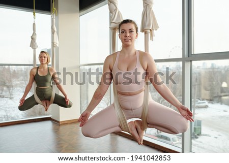 Two young energetic females in sportswear keeping balance while sitting in fly yoga hammocks during training with their feet put together