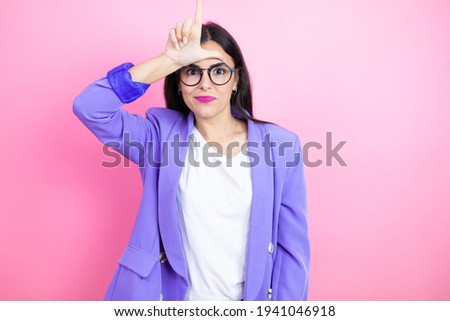 Young business woman wearing purple jacket over pink background making fun of people with fingers on forehead doing loser gesture mocking and insulting.