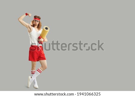 Full body funny goofy silly stupid skinny nerd in gym sweatband standing holding sports mat and dumbbells and showing weak bicep. Fitness workout advertising copyspace background, empty space for text