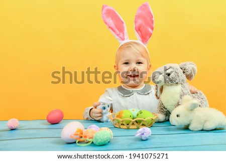 Little girl hugs and kisses Easter bunny on a yellow background. Painted eggs, soft toys for Easter.