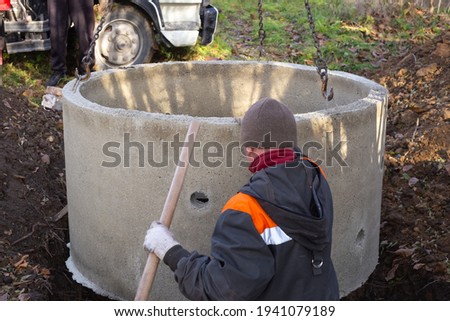 A loader lowers a concrete ring into a dug hole to build a septic tank. A worker installs a sewer into the ground. Royalty-Free Stock Photo #1941079189