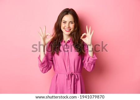 Beautiful woman 20s years, showing okay signs in approval, nod and smile pleased, standing satisfied, standing over pink background