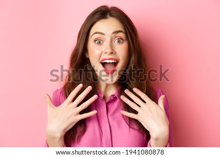 Close up of grateful and excited young woman gasping, holding hands on chest and stare with disbelief, receive awesome gift, standing against pink background