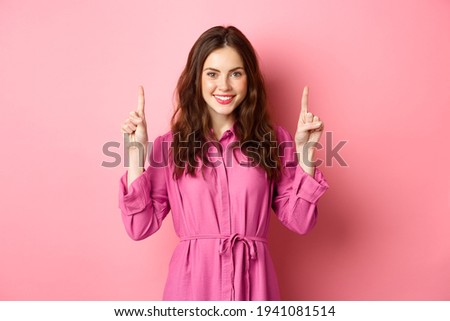 Stylish caucasian woman with curly hairdo and light trendy dress, smiling pleased, pointing fingers up at logo, showing copy space, standing against pink background