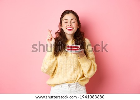 Celebration and holidays. Happy woman celebrates her birthday, makes wish with closed eyes and crossed fingers, holds bday cake with one candle, stands on pink background