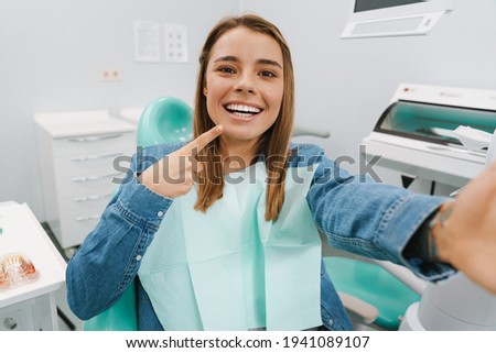 Joyful woman pointing finger at her smile while taking selfie in at dental clinic