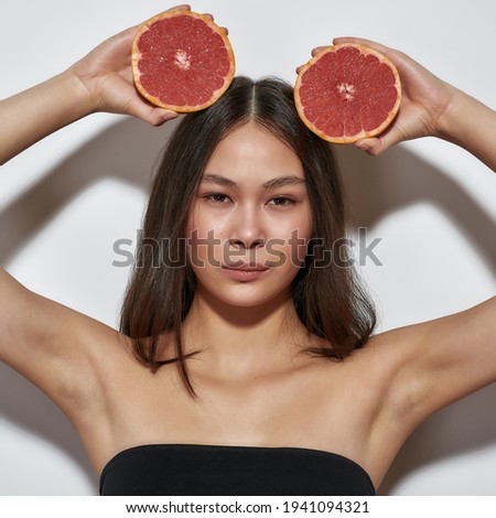 Beautiful girl standing in the studio with grapefruits. Young girl with clean skin on a light background. Fruit that helping with beauty concept. Brunette with grapefruit over her head