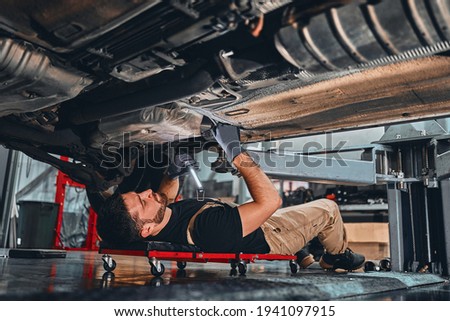 Young mechanic lying down and working under car at the garage. Repair undercarriage service. Selective focus. Royalty-Free Stock Photo #1941097915