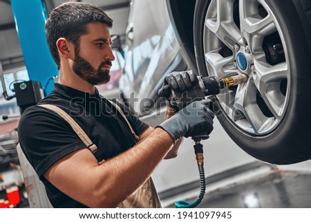 Car mechanic screwing or unscrewing car wheel of lifted automobile by pneumatic wrench at repair service station. Side view. Royalty-Free Stock Photo #1941097945