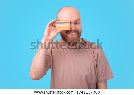 Charming handsome young bearded man holding credit card over eyes and smiling.