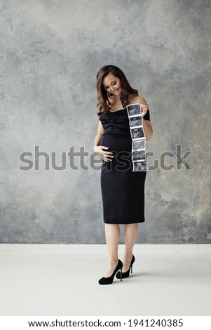 Pregnant woman looking at her tummy and holding ultrasound scan image on gray